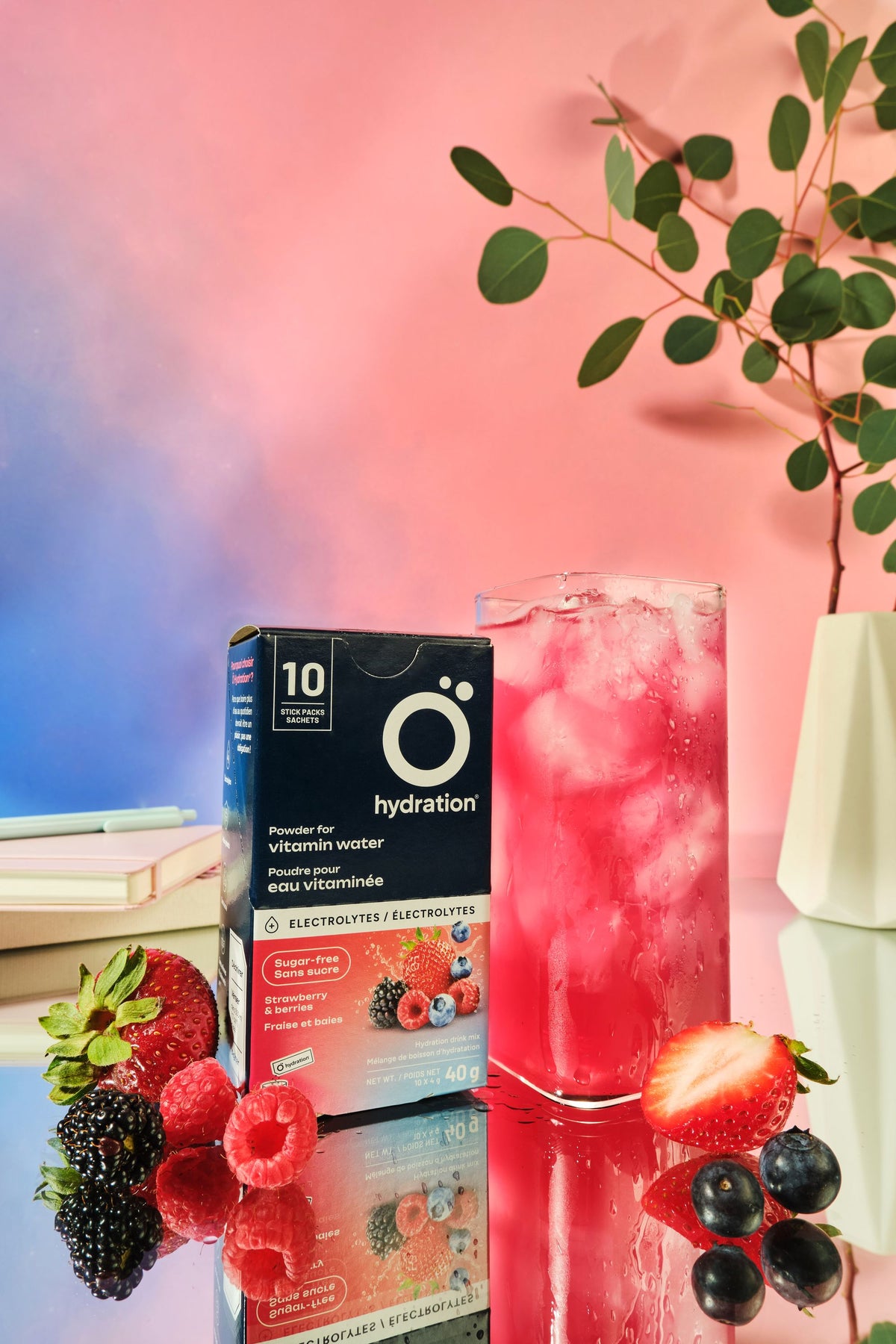 A box of O hydration electrolyte drink mix stands next to a tall glass of pink iced beverage with ice, styled with fresh strawberries, raspberries, blackberries, and blueberries on a reflective surface against a pink and blue gradient background.